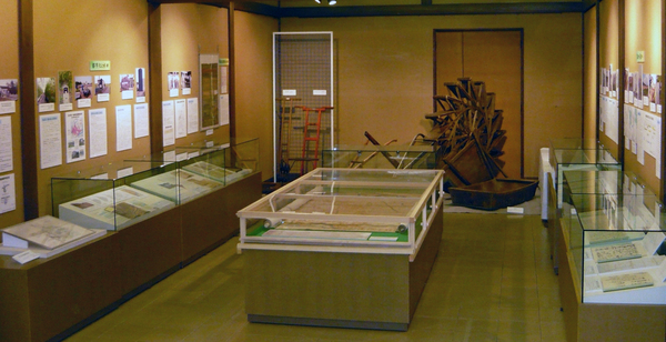 展示会場の両側に文書資料が入ったガラスケース、中央に図面入りの特大ケース、奥に農具が展示されています